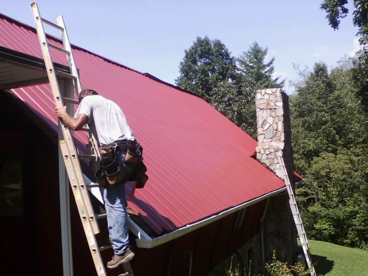 Roofer on a ladder beside a red metal roof and stone chimney — Soffit & Fascia Repair by Roofing Brothers in Halfway